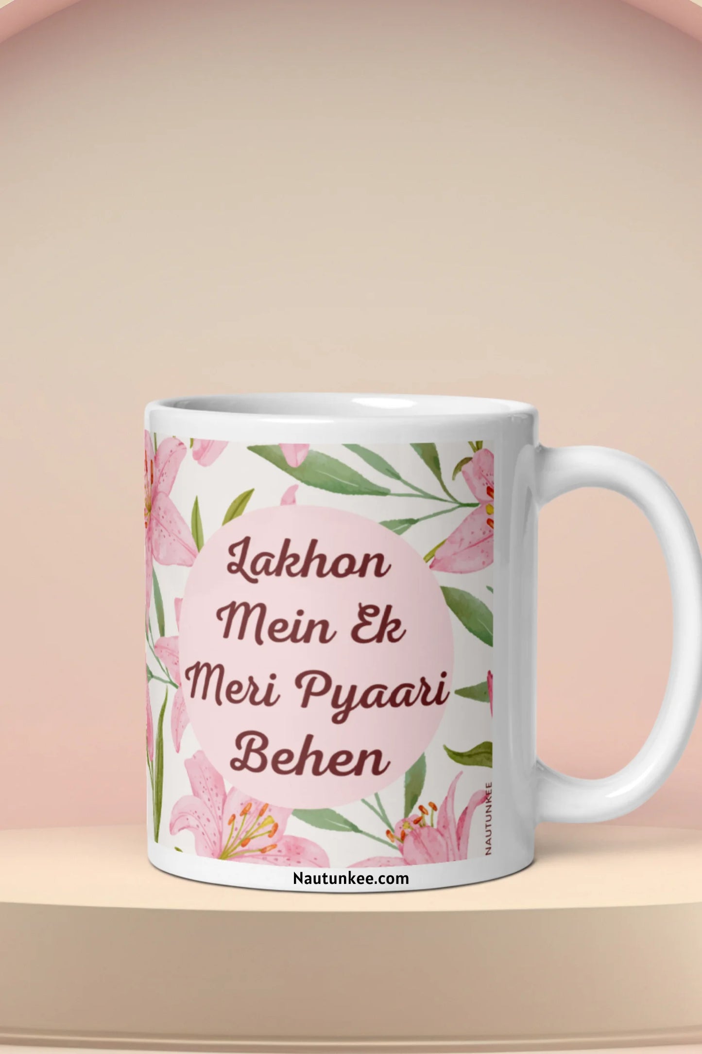 A ceramic mug for rakhi with a pink floral design and Hindi text reading 'Lakhon Mein Ek Meri Pyaari Behen' on a beige platform against a soft beige background.