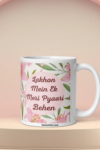 A ceramic mug for rakhi with a pink floral design and Hindi text reading 'Lakhon Mein Ek Meri Pyaari Behen' on a beige platform against a soft beige background.