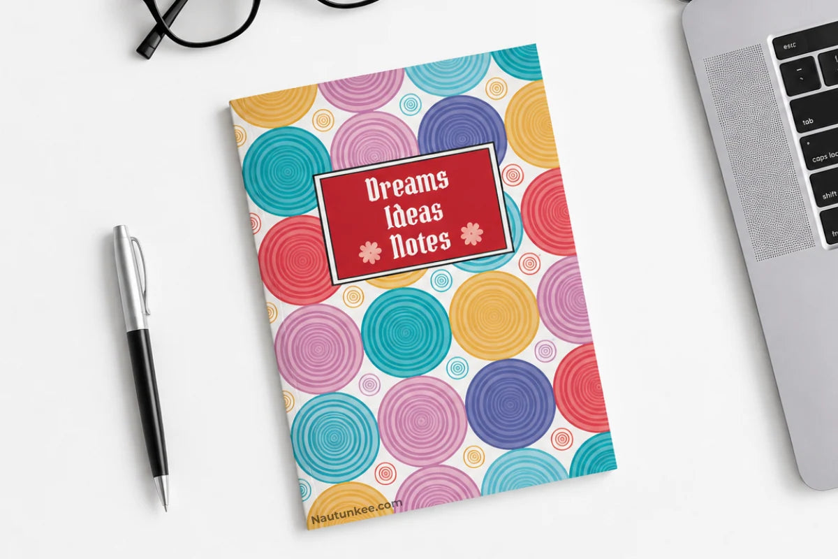A-5 notebook by Nautunkee titled 'Dreams Ideas Notes' with a colorful spiral pattern, styled on a white desk next to a pen, glasses, and a laptop.